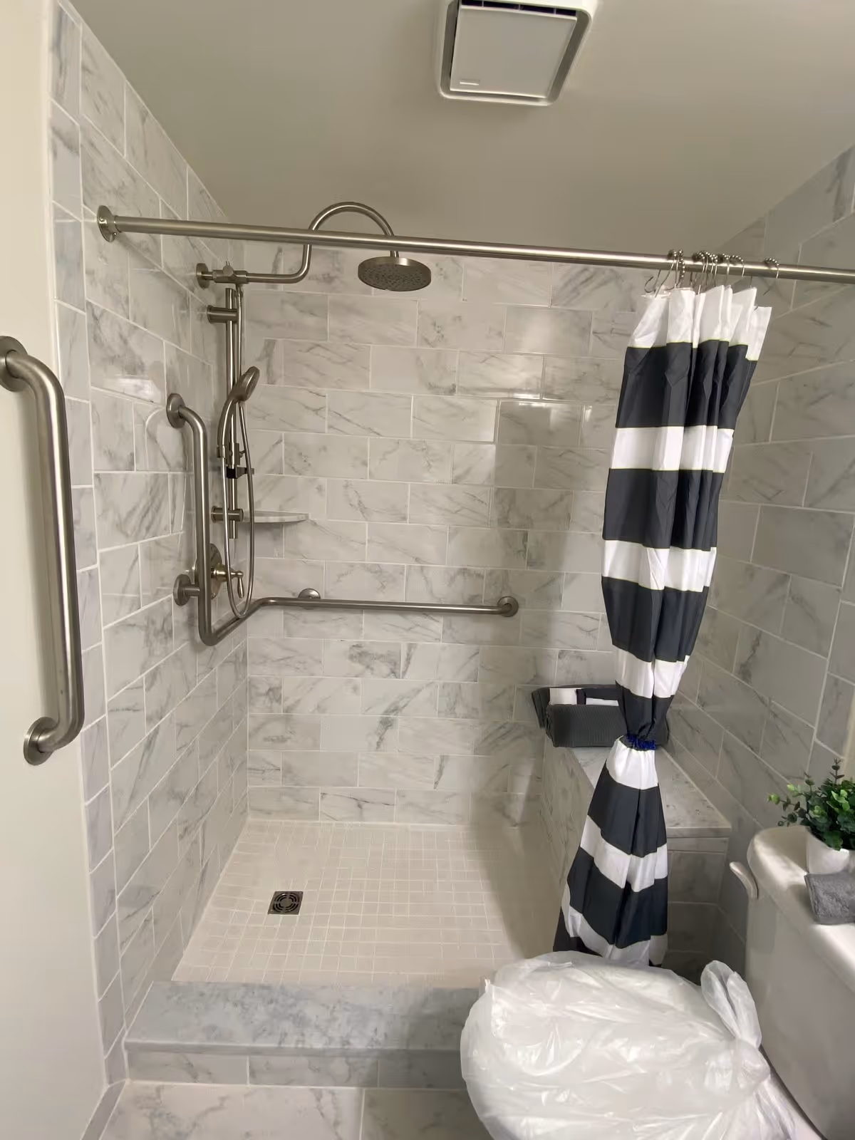 A modern bathroom shower area with white and gray marble tiles on the walls and floor. The shower has a stainless steel rain shower head, a handheld shower, and multiple grab bars for accessibility. A black and white striped shower curtain is tied back. There is a small bench inside the shower and a toilet with a plastic cover in the foreground. A small green plant is placed on the toilet tank.