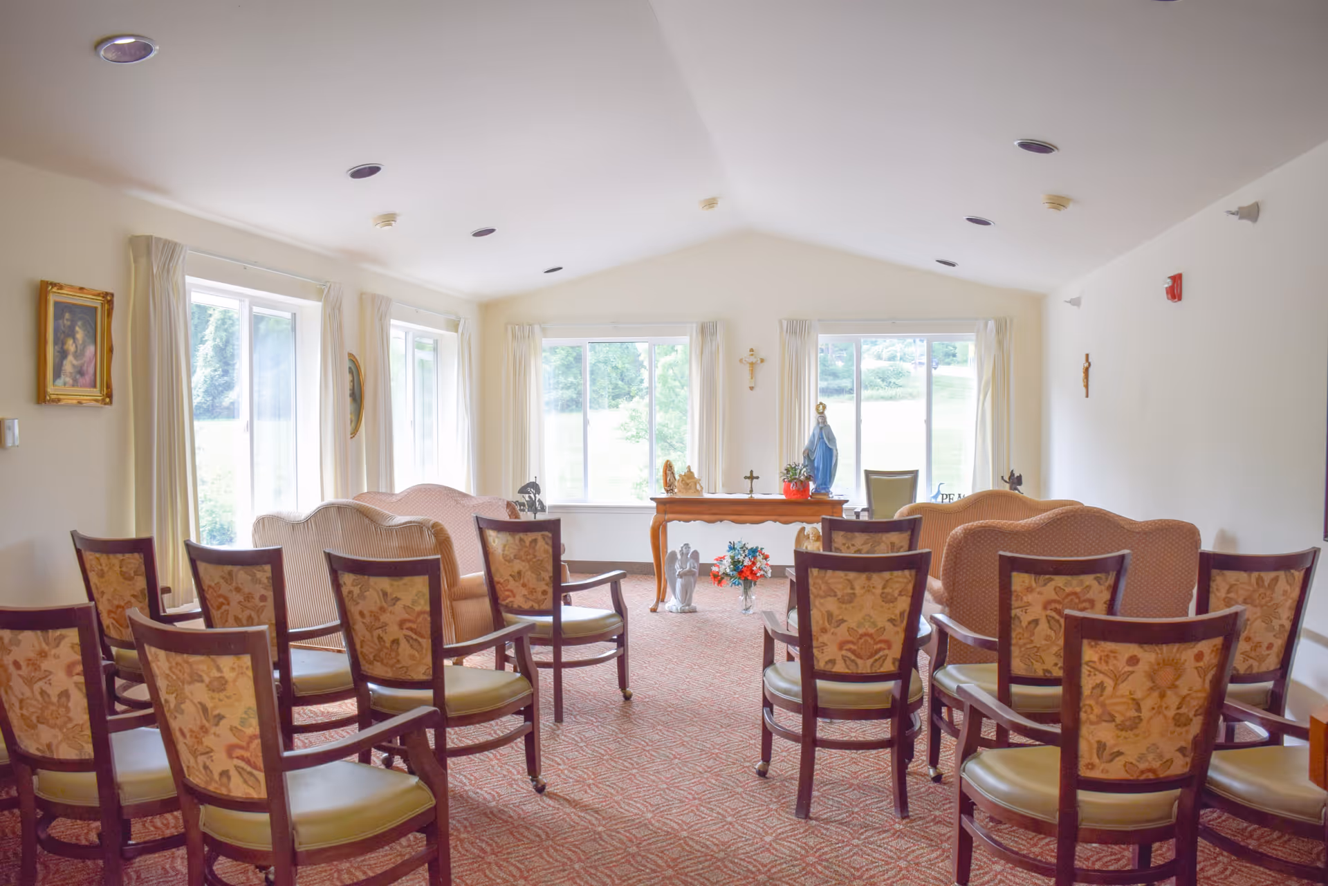 A bright room with multiple floral-patterned chairs arranged in rows facing a small altar table with religious statues and decorations. The room has large windows with cream-colored curtains letting in natural light, beige walls, and a carpeted floor. Religious artwork and crosses are displayed on the walls.