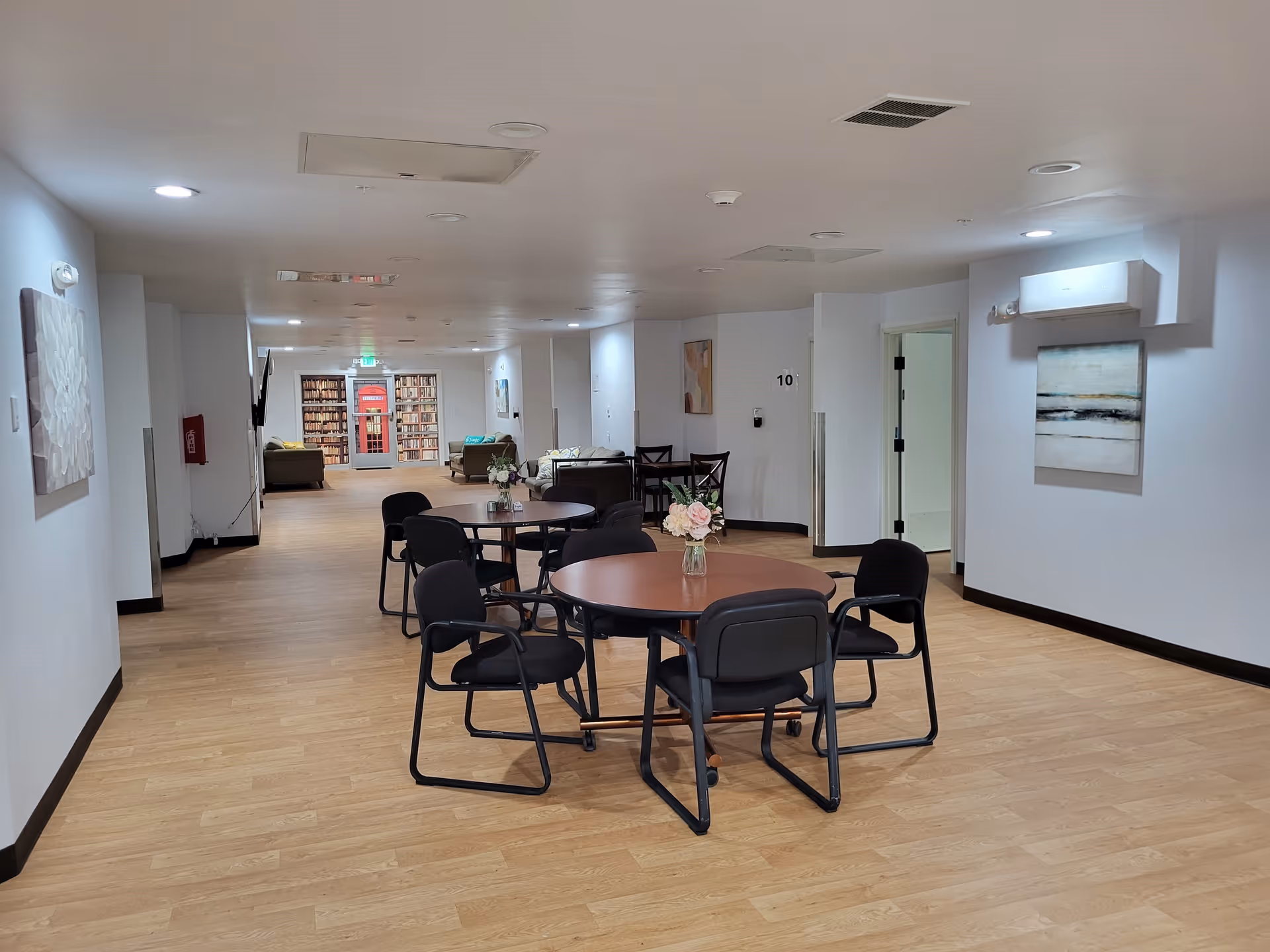 Spacious senior living common area with round tables and chairs, a seating area and bookshelf visible in the background.
