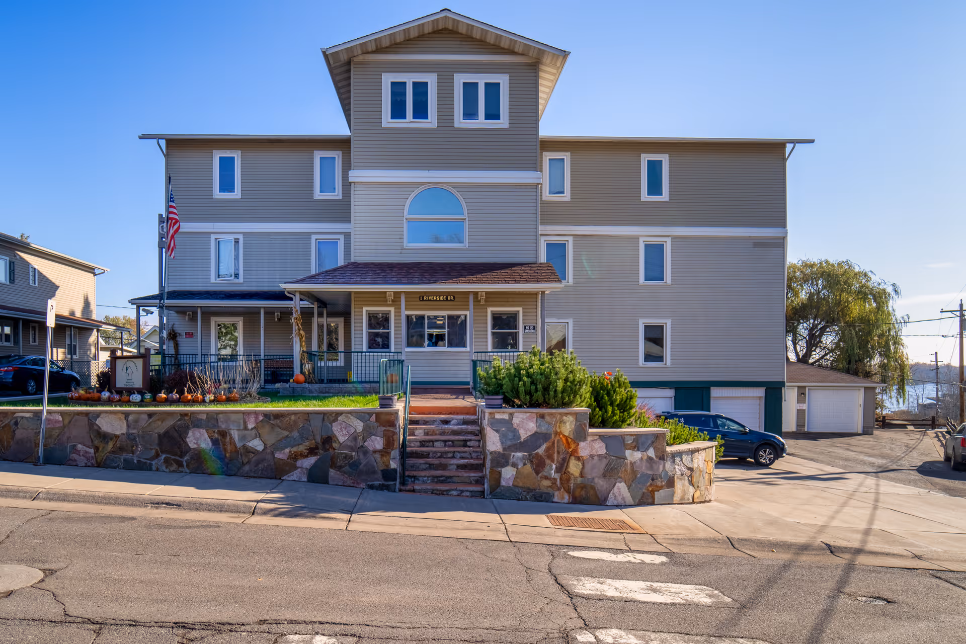 Front exterior of a three-story senior living building with stone retaining wall, steps, landscaping and an American flag.