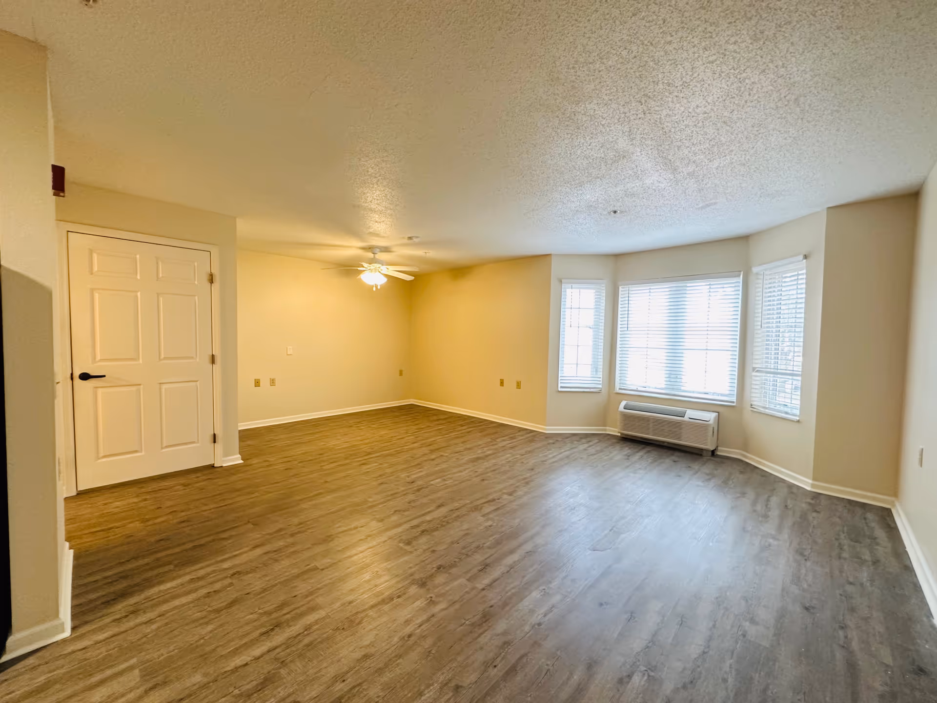 Spacious empty living room with wood-look flooring, a ceiling fan, bay windows, and a wall air unit.