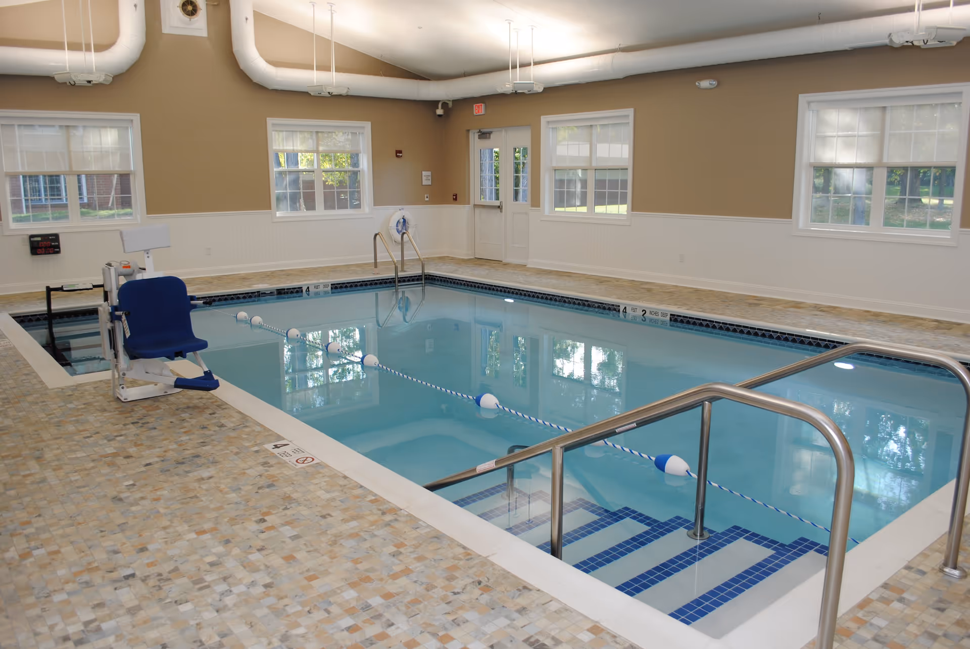 Indoor swimming pool with tiled floor, metal handrails, and a blue pool lift chair for accessibility. The room has beige walls, several windows letting in natural light, and white ceiling pipes.