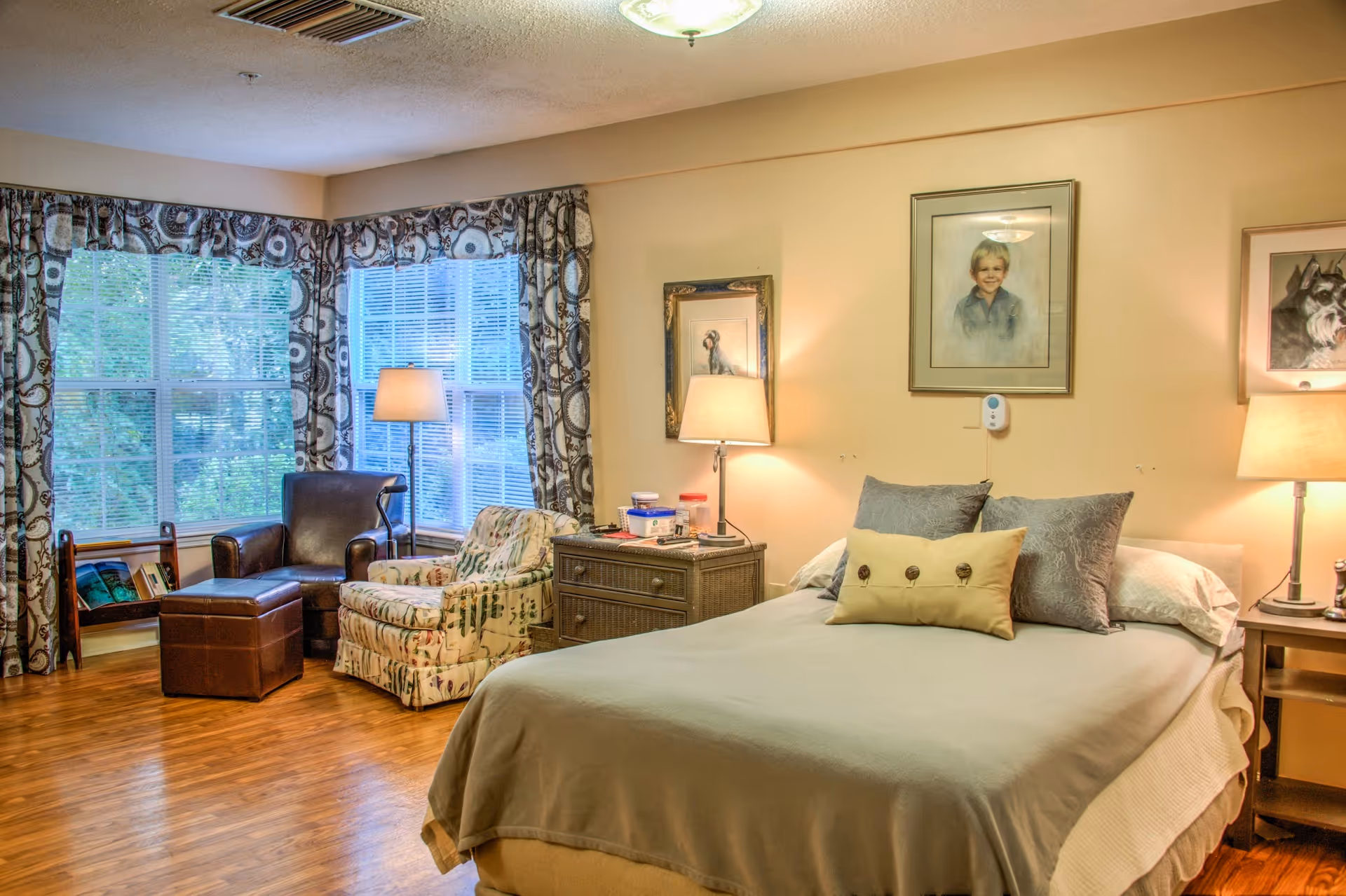 A cozy bedroom with a neatly made bed featuring multiple pillows, two bedside tables with lamps, and framed pictures on the wall above the bed. There is a sitting area with a leather armchair, an upholstered armchair, and a small ottoman near large windows with patterned curtains. The room has wooden flooring and soft lighting.