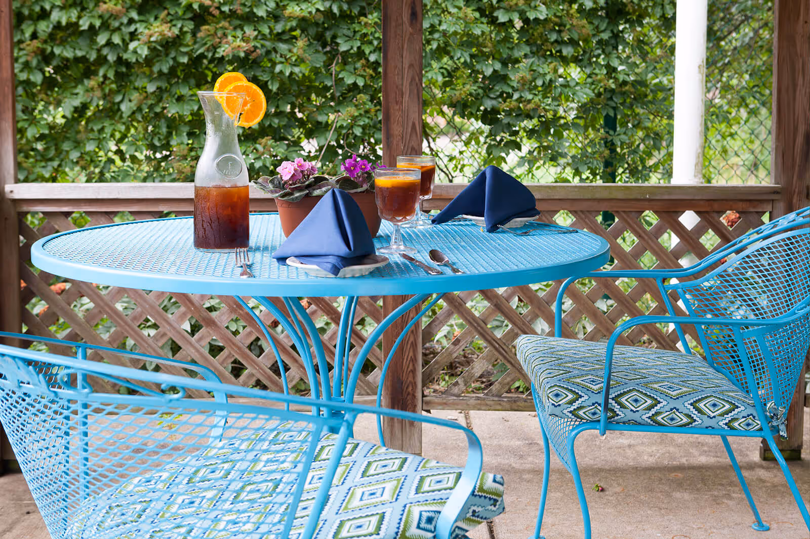 Blue metal patio table and chairs on a covered outdoor patio with a pitcher and glasses of iced tea, napkins, and a small potted flower.