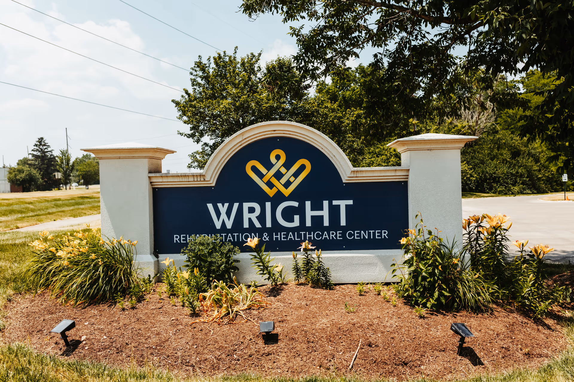 Outdoor sign for Wright Rehabilitation and Healthcare Center surrounded by landscaping with flowers and greenery, set against a backdrop of trees and a partly cloudy sky.