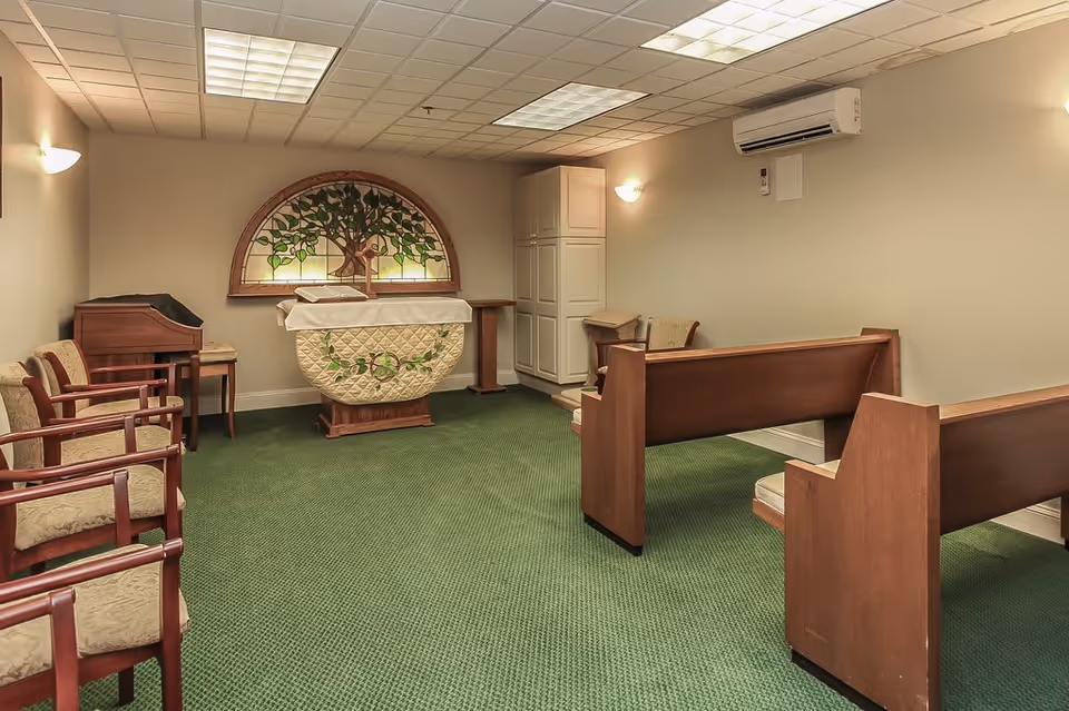 Small chapel room with green carpet, wooden pews, chairs, a lectern covered with a decorative cloth, and a stained glass window featuring a tree design on the back wall. The room has beige walls, ceiling lights, and an air conditioning unit mounted on the wall.