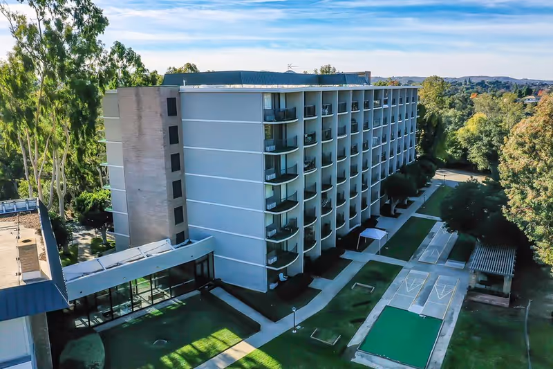 Exterior view of a multi-story senior living facility building with balconies, surrounded by trees and greenery. There are outdoor shuffleboard courts and walking paths in the foreground.