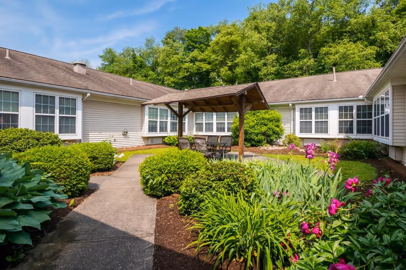 Outdoor courtyard area at Celebration Villa of Loyalsock featuring a paved walkway, green bushes, colorful flowers, a wooden pergola with a table and chairs underneath, and surrounded by a single-story building with multiple windows and a backdrop of tall green trees.