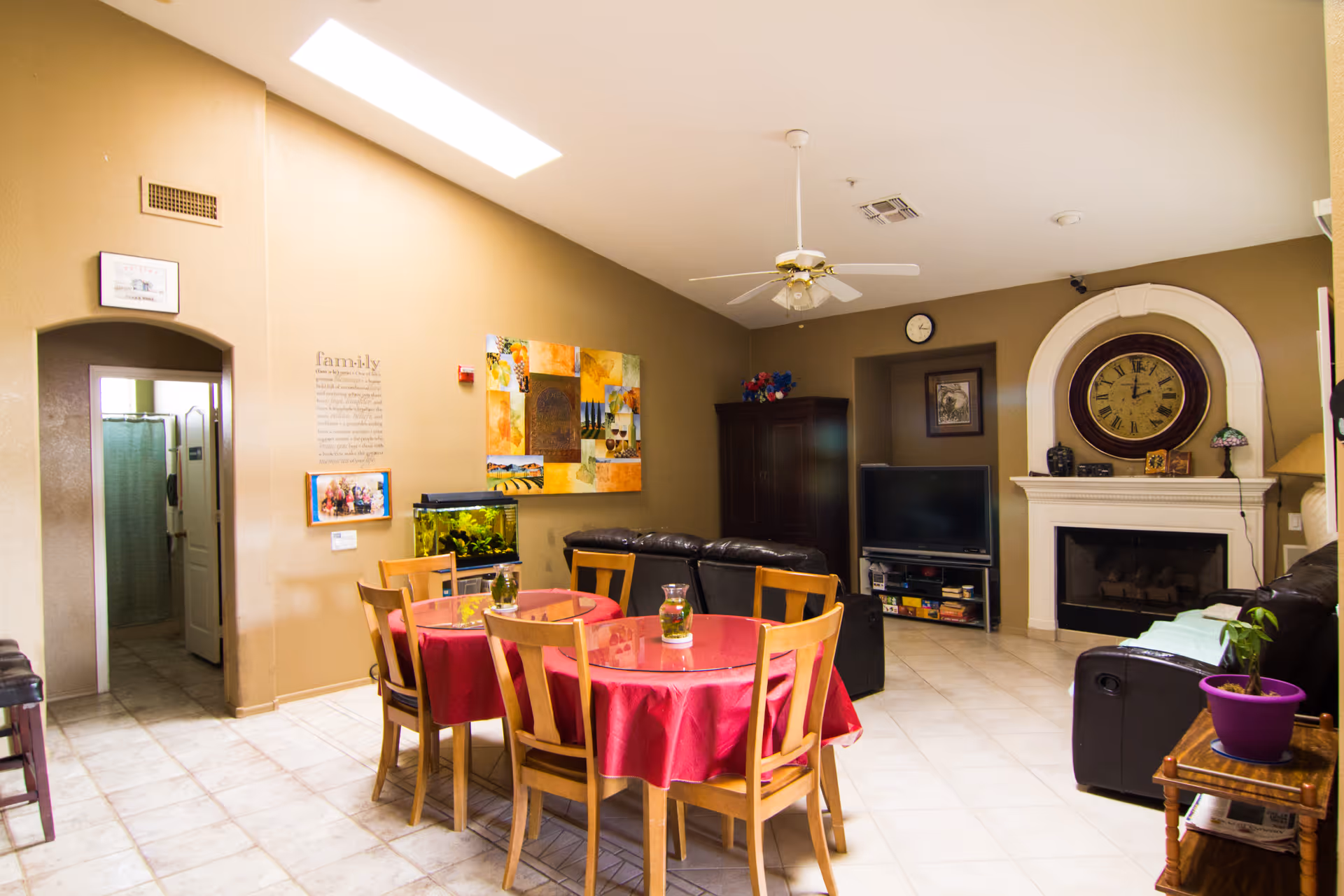 A cozy living room and dining area in an assisted living home. The dining area has a round table covered with a red tablecloth and surrounded by wooden chairs. The living room features a black leather couch, a large wall clock above a white fireplace, a flat-screen TV on a stand, and a ceiling fan. The walls are painted beige and decorated with colorful artwork and a family-themed wall decal. There is also a fish tank near the dining table and a small wooden side table with a purple potted plant.