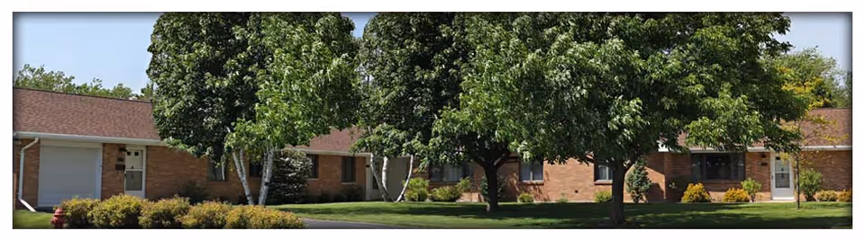 One-story brick senior living building front with large trees, a green lawn, and a driveway.