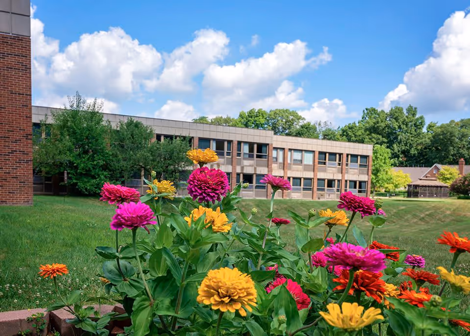 Colorful flowers in the foreground with a two-story brick and beige building in the background under a blue sky with scattered clouds.
