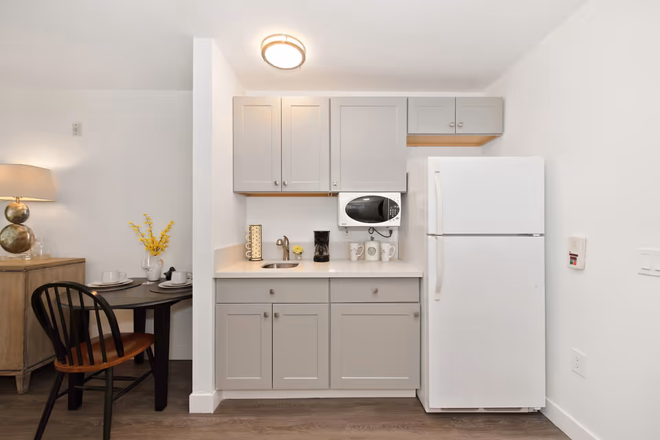 Small kitchenette with gray cabinets, a white refrigerator and microwave, a sink and coffee maker, and a nearby small dining table with chairs.