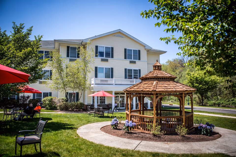 Front of a three-story assisted living building with a wooden gazebo on a grassy courtyard and tables with red umbrellas.