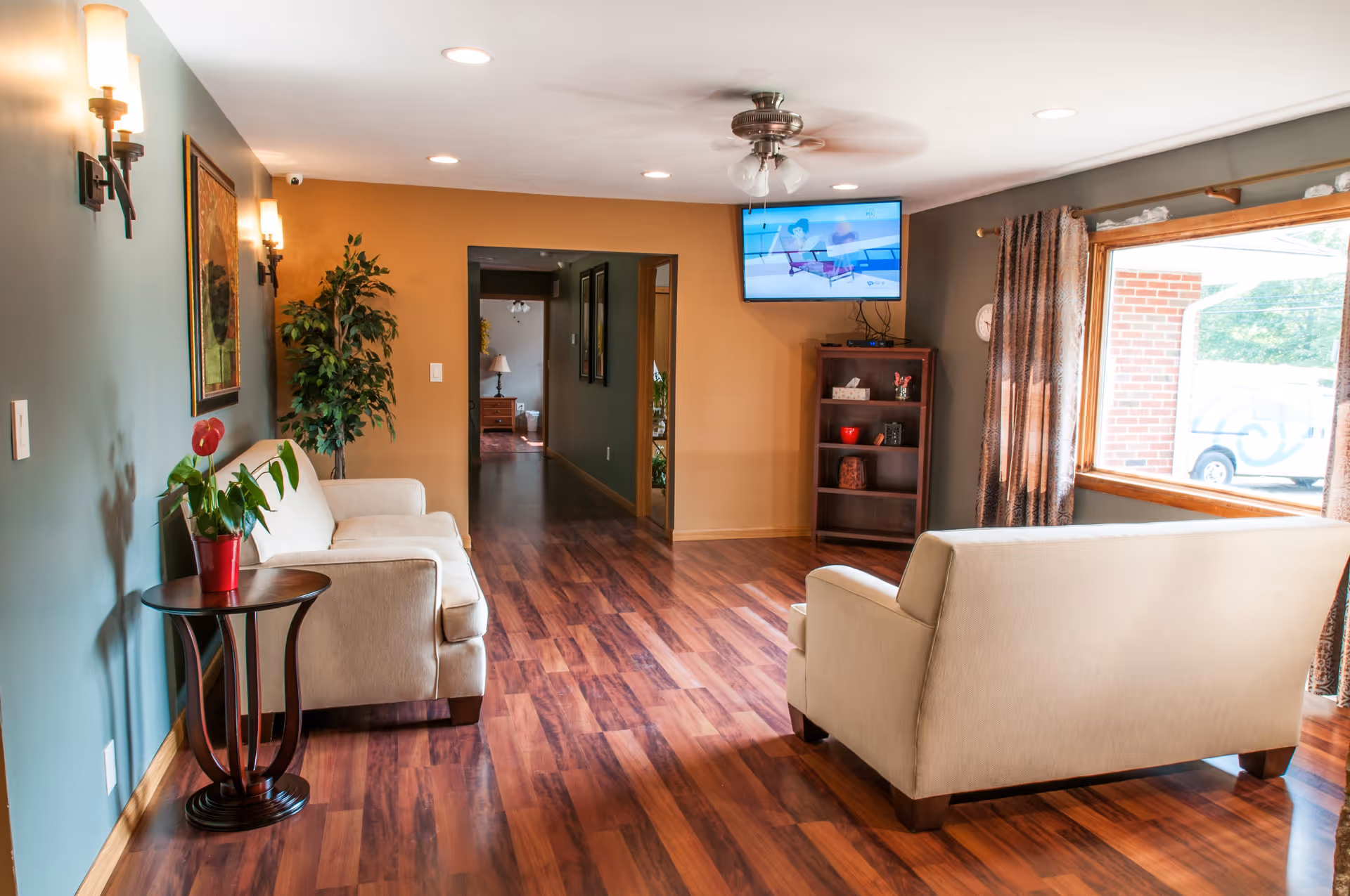 A cozy living room area with two beige sofas facing each other on a wooden floor. There is a small round wooden side table with a potted plant on it next to one sofa. The walls are painted in two colors, teal and mustard yellow. A flat-screen TV is mounted on the mustard yellow wall above a wooden bookshelf with decorative items. A large window with patterned curtains lets in natural light, and a ceiling fan with lights is on the white ceiling.