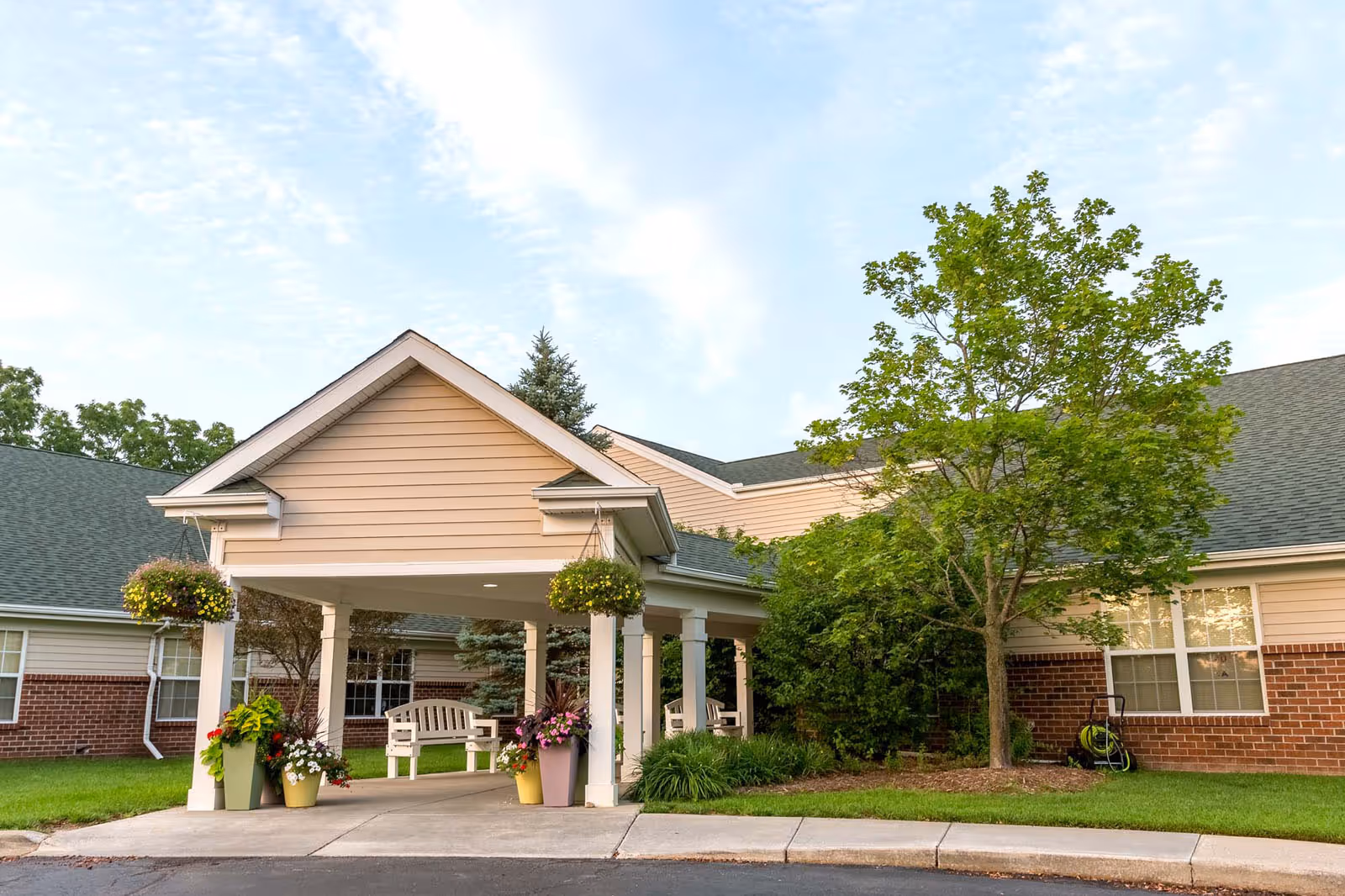 Covered portico entrance of a brick-and-siding senior living building with hanging baskets, potted flowers, and a bench.