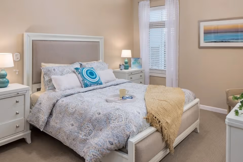 A cozy bedroom with a large bed featuring a patterned comforter, multiple pillows including a decorative blue one, and a beige throw blanket. The room has two white nightstands with lamps, a window with white curtains, a framed picture on the wall, and a beige armchair partially visible.