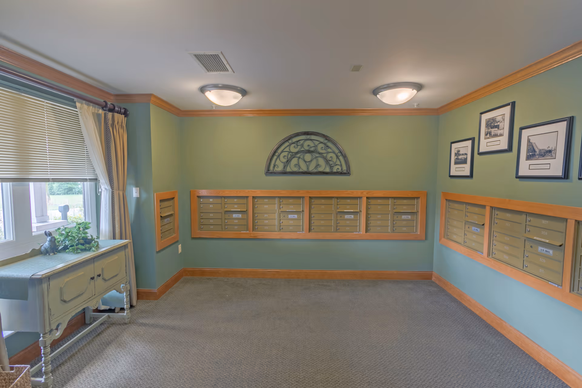 A small room with green walls and beige carpet featuring multiple mailboxes mounted on the walls. There is a decorative metal wall art piece above the mailboxes on the center wall. A window with blinds and curtains is on the left side, next to a small table with a plant and a decorative item on it. Three framed black and white pictures hang on the right wall above the mailboxes.
