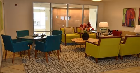A cozy living room area with modern furniture including two green armchairs, a green loveseat, and four blue chairs around a small round table. There is a wooden coffee table with a decorative vase and flowers in the center. The room has a large window with a view of an exterior wall, a floor lamp, and colorful artwork on the walls.