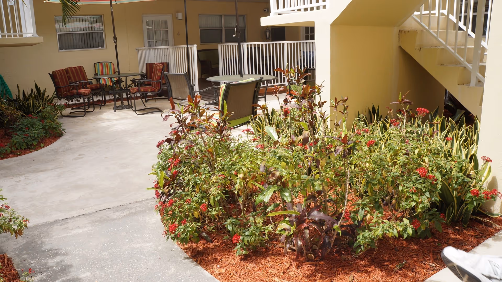 Outdoor courtyard with patio tables and chairs, a planted flower bed, and surrounding apartment doors and stairway.