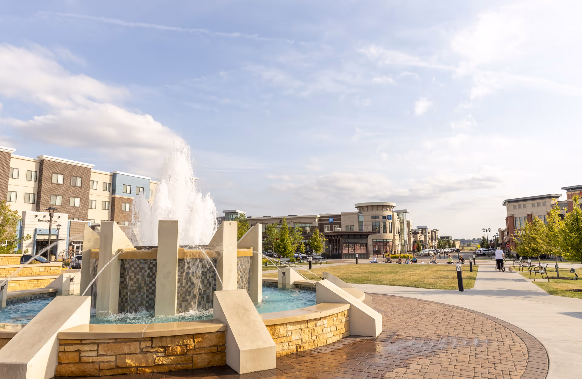 Outdoor plaza area with a large decorative water fountain in the foreground, surrounded by paved walkways, benches, and green lawn areas. Modern multi-story buildings and shops are visible in the background under a partly cloudy sky.