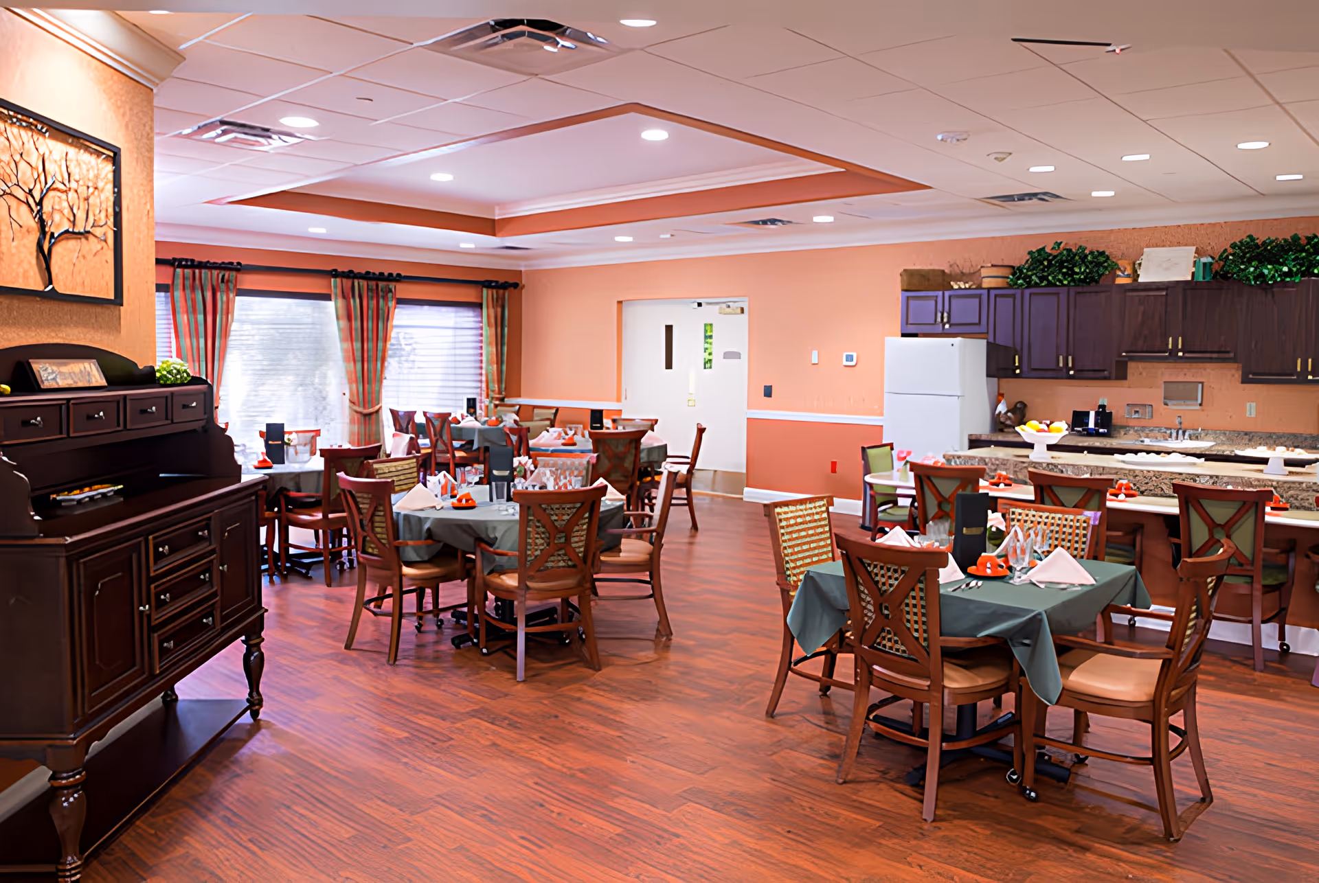 A cozy dining room with several tables covered in green tablecloths, each set with folded napkins, cups, and utensils. The room has wooden chairs, a wooden floor, and a kitchen area with dark cabinets, a white refrigerator, and a countertop with bar stools. The walls are painted in warm tones with windows covered by striped curtains, and there is a decorative wooden cabinet on the left side.