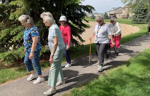 Five elderly women walking outdoors on a paved path surrounded by grass and trees. Two of the women are using walking aids, and the weather appears sunny and pleasant.