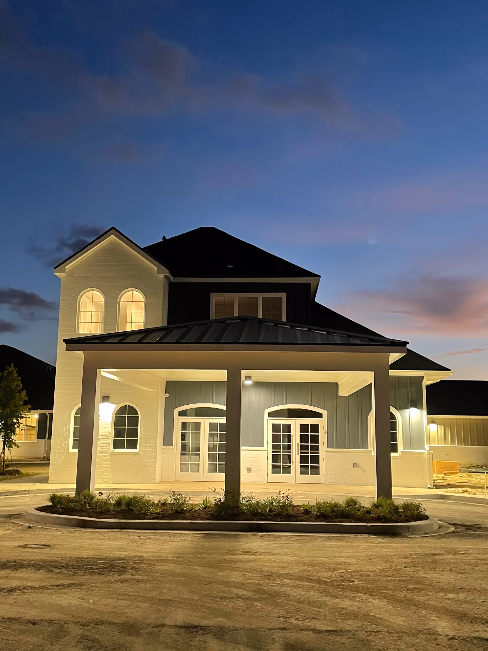 Illuminated front entrance of a two-story building at dusk with a covered portico, arched windows, and double French doors.