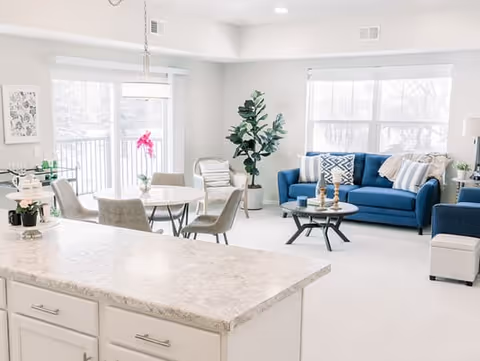 Bright open-concept living area with a kitchen island in the foreground, dining table, blue sofa, armchairs, and large windows.