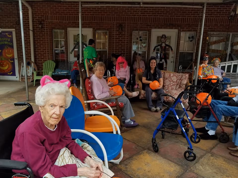 A group of elderly people and a caregiver sitting outside on a covered patio decorated for Halloween. Some individuals hold orange pumpkin buckets, and one elderly woman in the foreground wears pink cat ears. The background shows brick walls, windows, and Halloween decorations including a poster with pumpkins and a figure on a door.