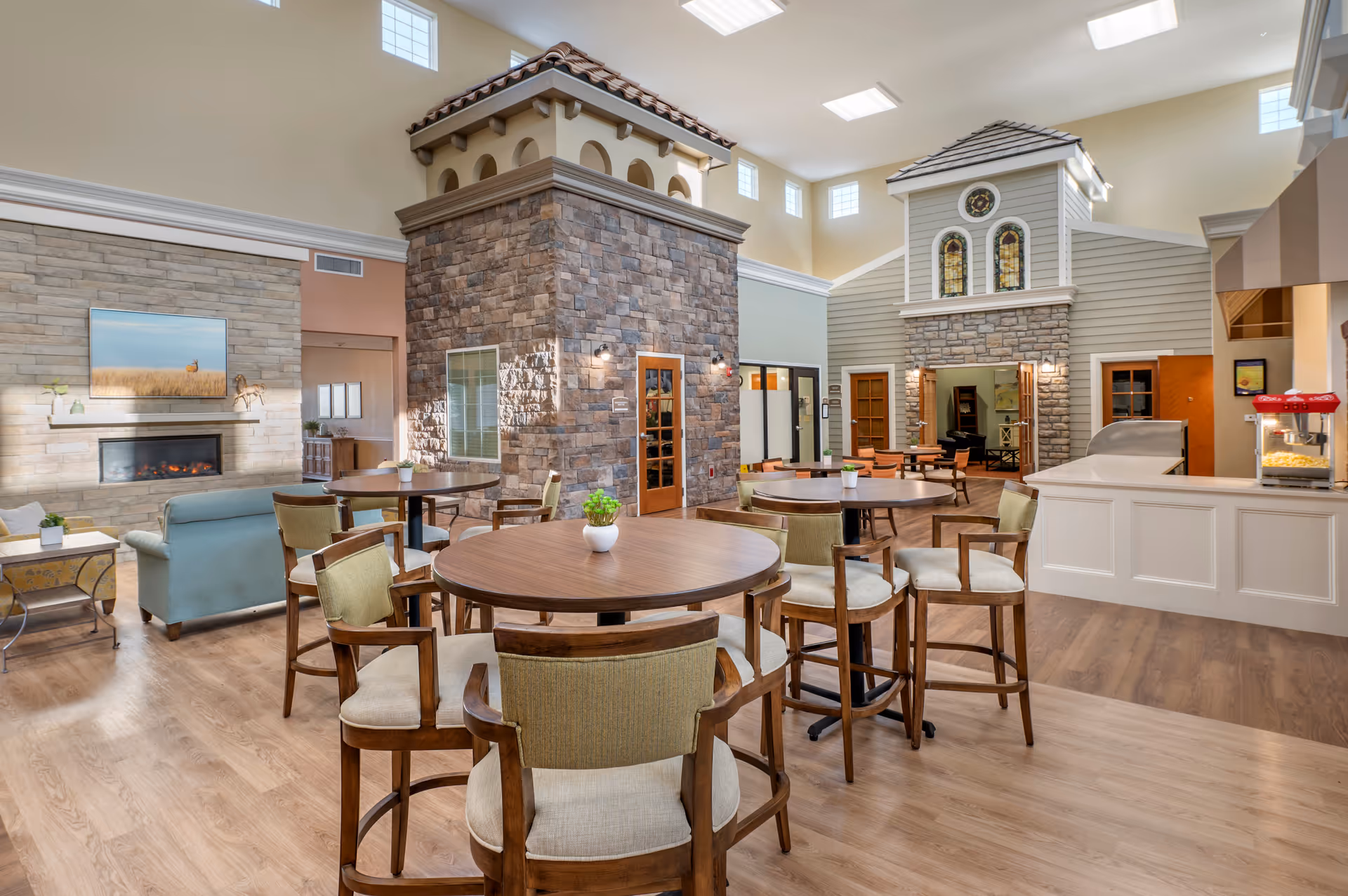 Bright, village-style senior living common area with round dining tables and chairs, lounge seating, a fireplace, and a small serving counter.