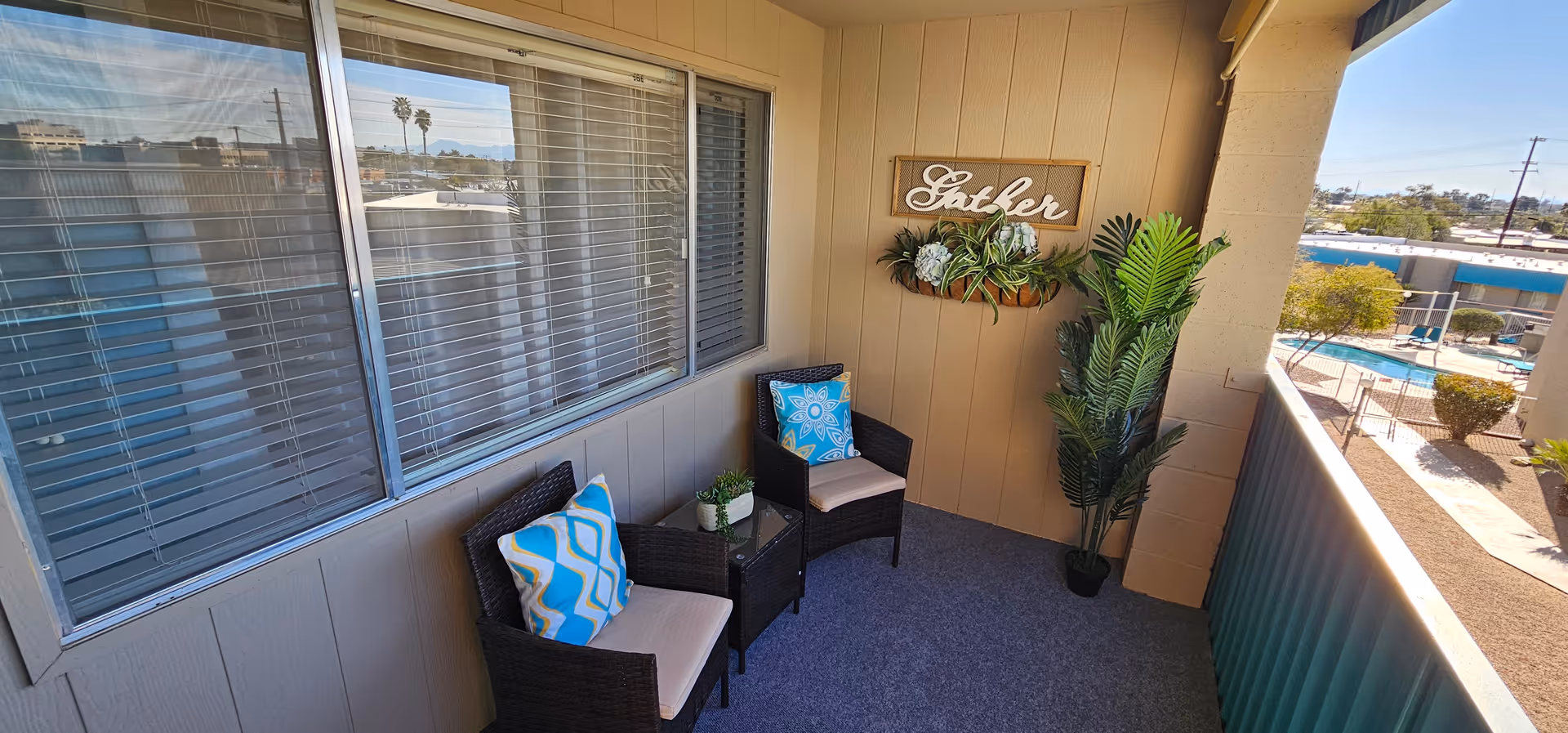 A small outdoor balcony area with two wicker chairs featuring blue and white patterned cushions, a small glass-top table with a plant, a large potted plant in the corner, and a decorative wall hanging with the word 'Gather' above a planter with greenery. The balcony overlooks a pool area and surrounding buildings under a clear blue sky.