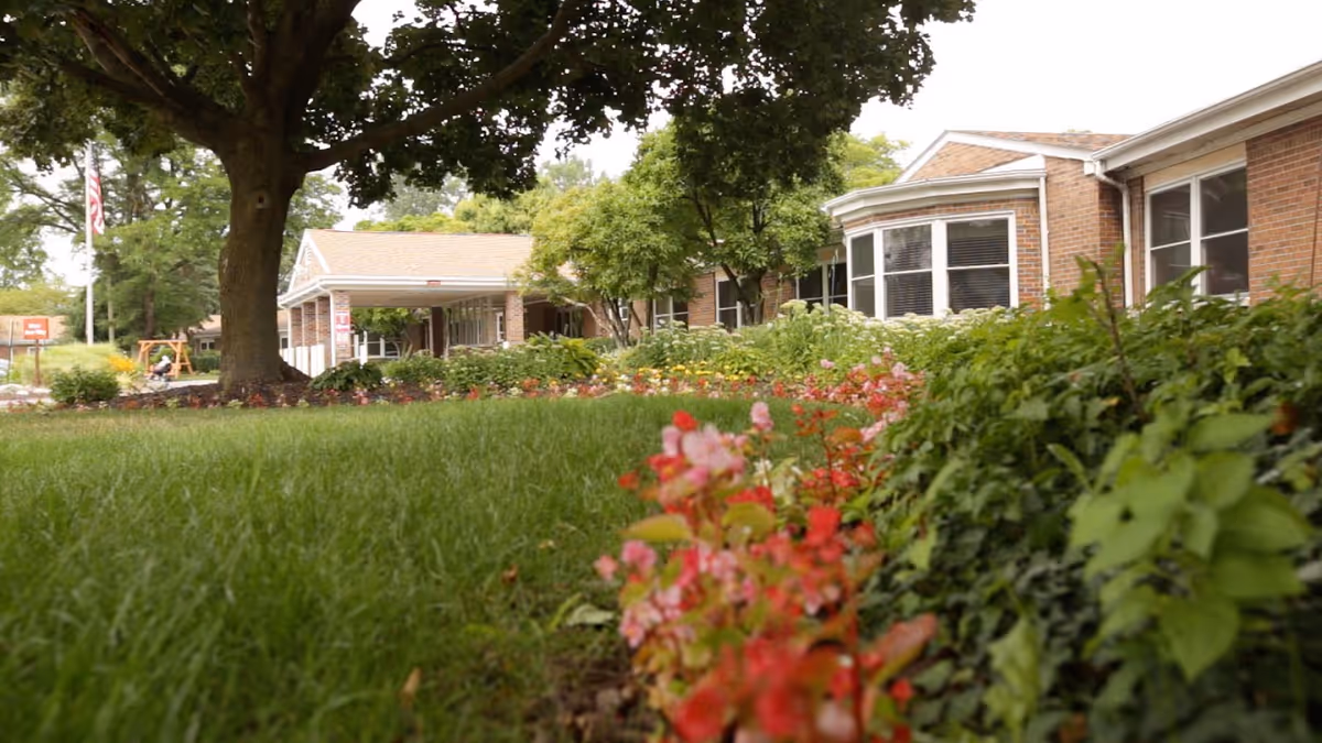 View of a senior living facility's outdoor garden area with green grass, colorful flowers, trees, and a brick building with large windows in the background.