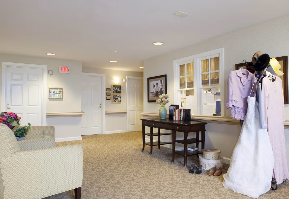 A well-lit hallway in a senior living facility with beige patterned carpet and light-colored walls. There are three white doors, an exit sign, framed pictures on the walls, and a wooden table with books and a vase of flowers. To the right, a clothing rack holds a white wedding dress, a lavender suit jacket, and a pink dress, along with hats and shoes on the floor. Two upholstered armchairs with floral arrangements are partially visible on the left.