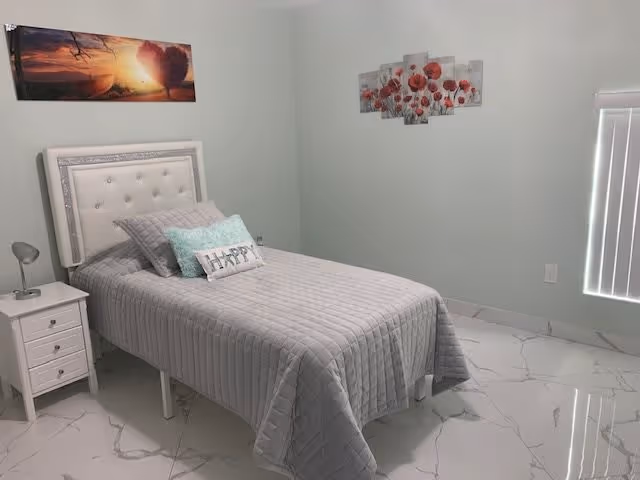 A neatly made single bed with a tufted white headboard, gray quilted bedspread, and decorative pillows including one with the word 'HAPPY'. The bed is positioned next to a white nightstand with a small lamp. The room has light-colored walls with two pieces of wall art: one depicting a sunset with a heart-shaped tree and another featuring red flowers. The floor is covered with white marble tiles, and there is a window with vertical blinds partially visible on the right side.