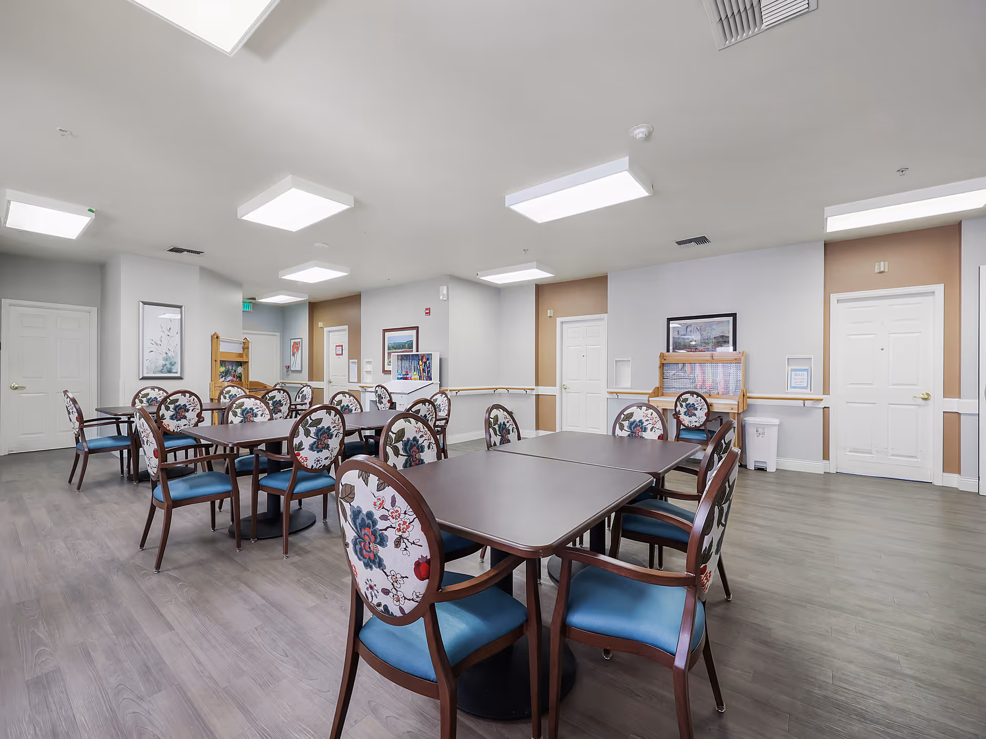 Communal dining room with dark tables and floral-upholstered chairs in a bright senior living common area.