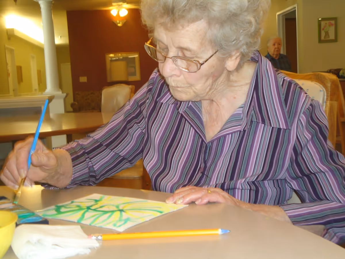 An elderly woman wearing glasses and a purple striped shirt is painting with a brush on a piece of paper at a table in a well-lit room. Another elderly person is visible in the background, sitting near a doorway. The room has warm lighting and comfortable furniture.