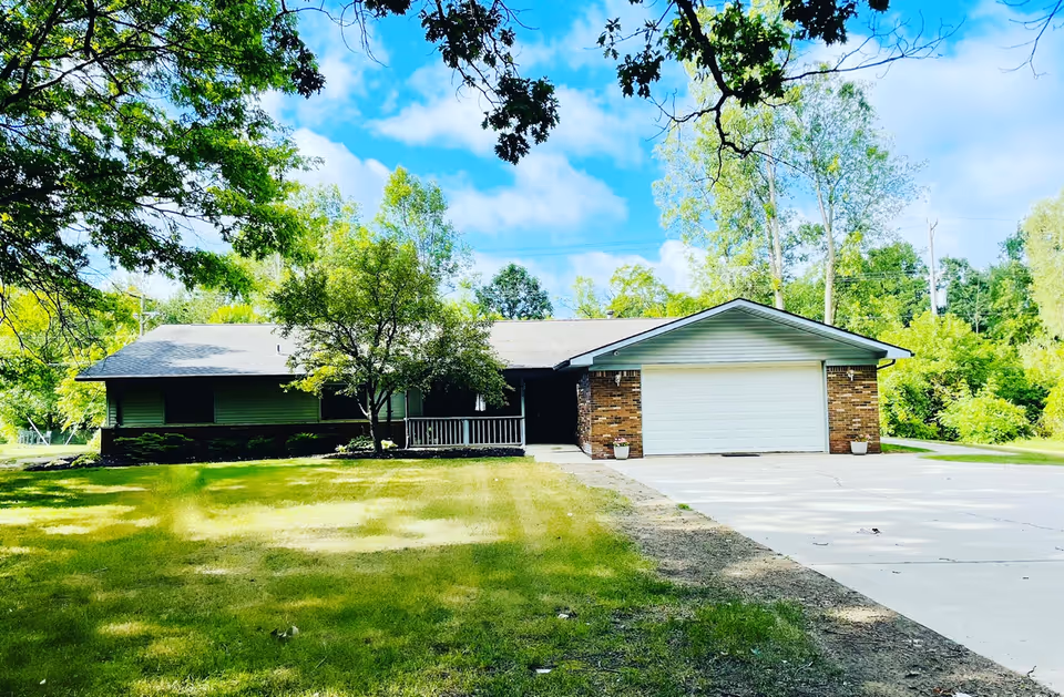 Single-story brick house with a large front lawn, a concrete driveway leading to a two-car garage, and trees surrounding the property under a partly cloudy blue sky.