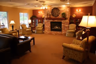 A cozy living room with multiple armchairs and sofas arranged around a brick fireplace. The room is warmly lit with table lamps and wall sconces, and has large windows letting in natural light. There is a decorative circular piece above the fireplace and a wooden cabinet with plants and decor items.