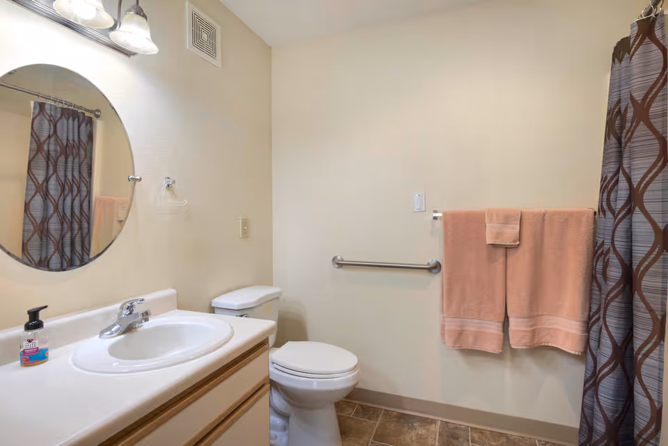 A clean bathroom with a white sink and countertop, a round mirror above the sink, a toilet next to the sink, a towel rack with two peach-colored towels, and a shower curtain with a brown and gray geometric pattern.