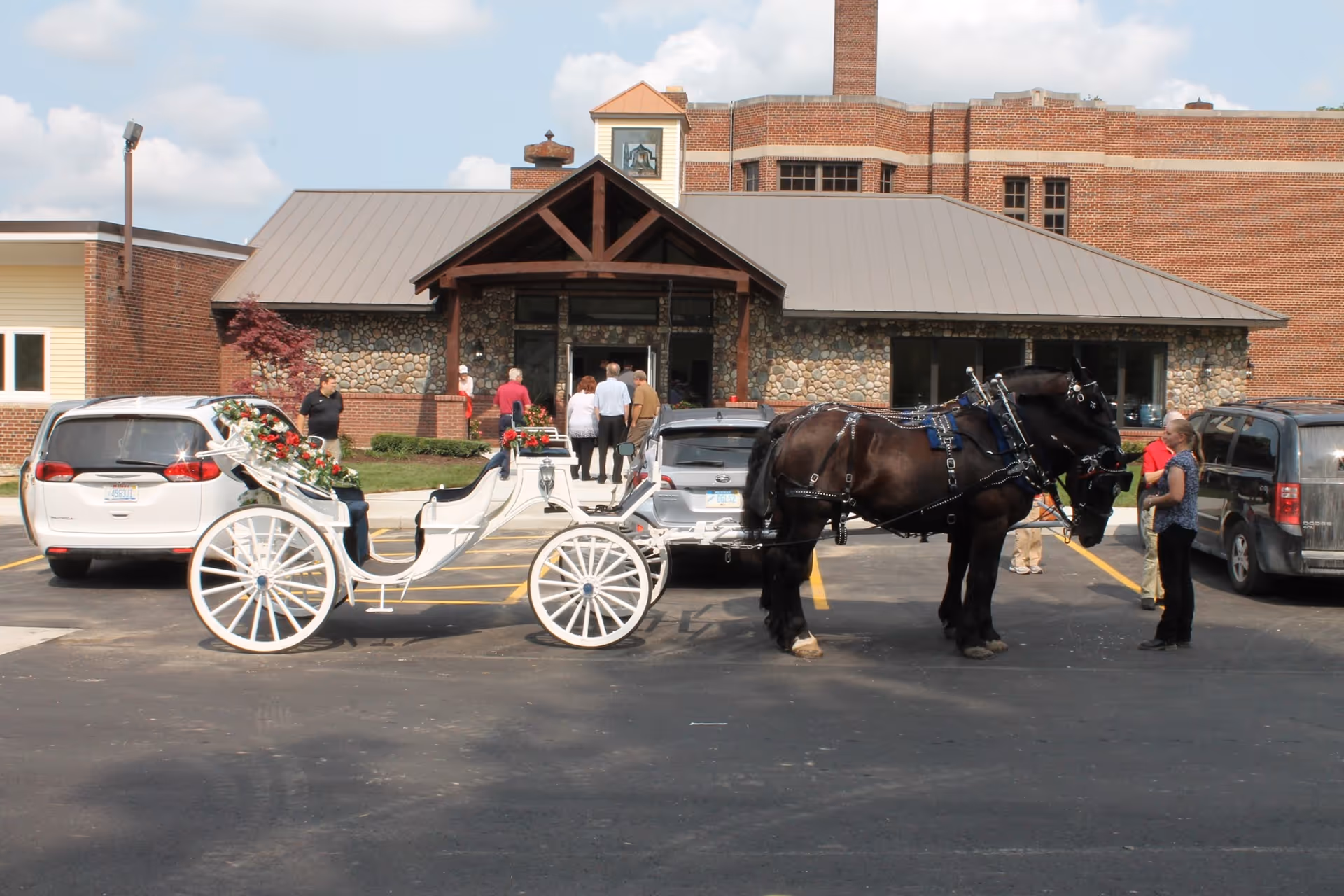 A white horse-drawn carriage hitched to a black horse parked in front of the brick entrance to a senior apartment building with people nearby.