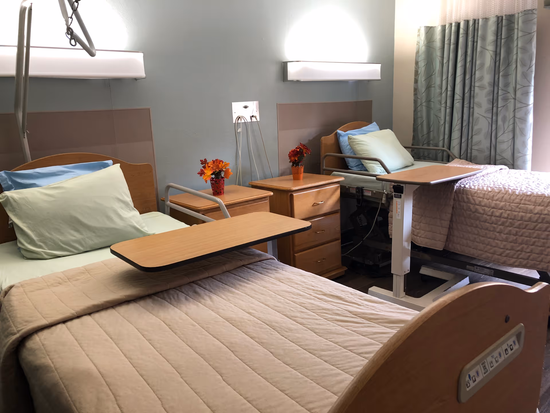 A room with two hospital-style beds, each with beige quilts, light green pillows, and overbed tables. Between the beds are two wooden nightstands with small flower arrangements. The room has light blue walls and a window with patterned curtains.