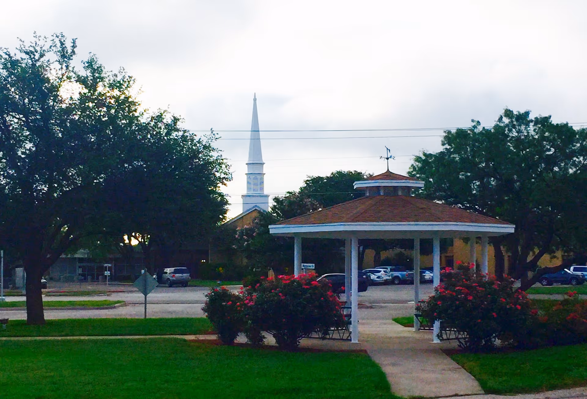 Outdoor view of a gazebo surrounded by bushes with red flowers, green grass, and trees. In the background, there is a parking lot with several cars and a white church steeple visible behind the trees.