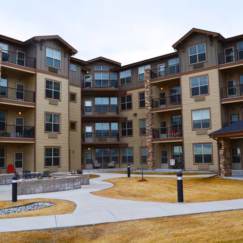Exterior view of a multi-story senior living facility building with balconies, large windows, and a courtyard featuring paved walkways, grass, and outdoor seating areas.