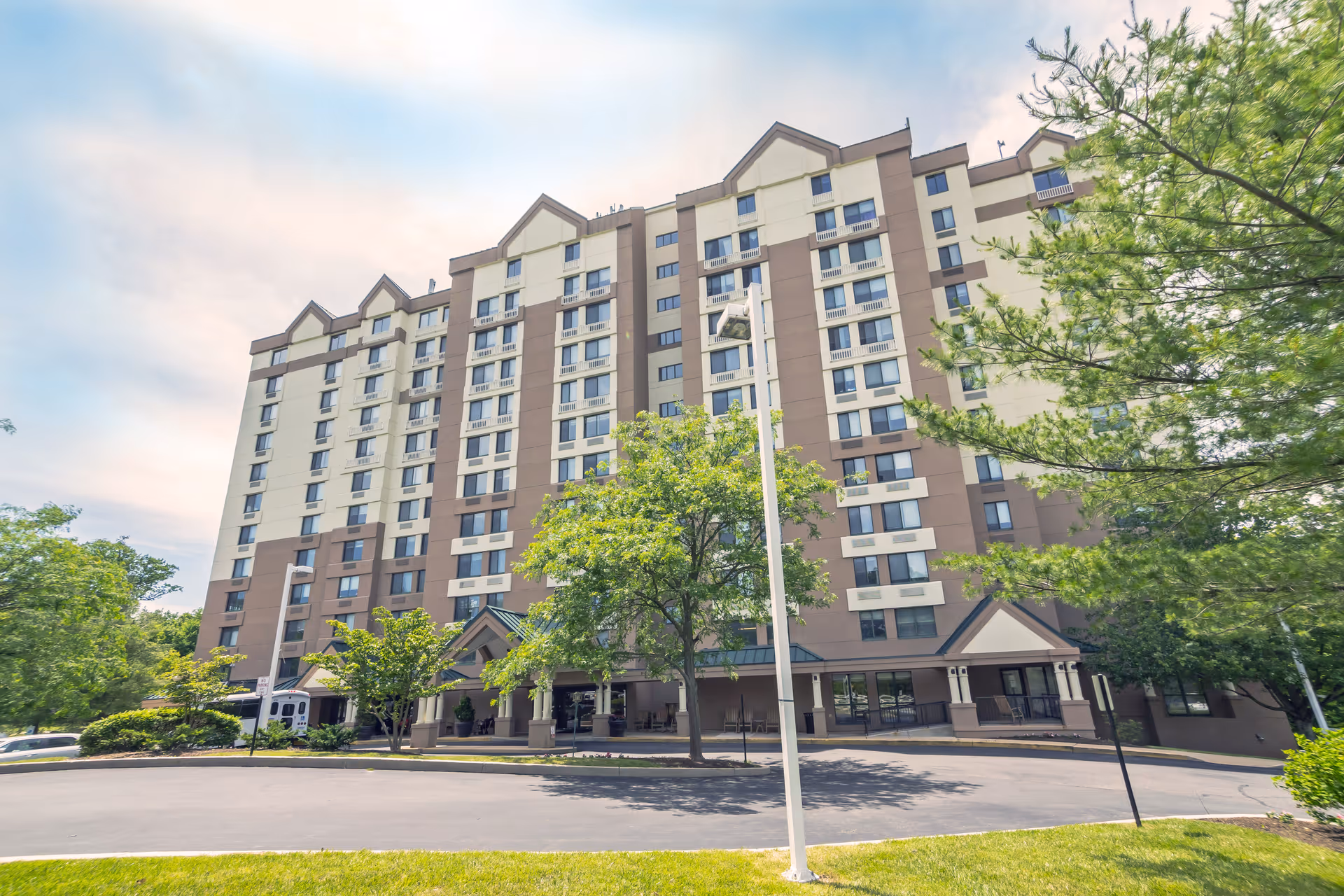 Front exterior of a multi-story beige-and-brown senior living building with trees, a circular driveway, and main entrance.