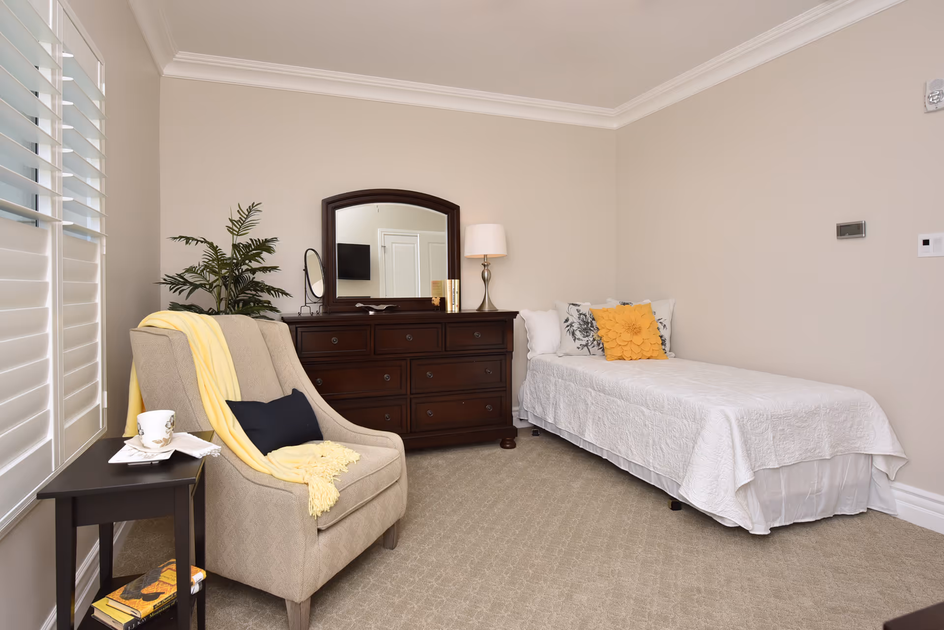 A cozy bedroom with a single bed covered in a white quilt and decorated with floral and yellow pillows. Next to the bed is a dark wooden dresser with a mirror, a table lamp, and some books. A beige armchair with a yellow throw blanket and a black pillow is positioned near a small side table holding a cup and a napkin. A green potted plant is placed behind the armchair. The room has beige walls, carpeted floor, and white plantation shutters on the window.