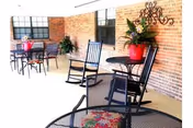 Outdoor patio area with black metal tables and chairs, including two rocking chairs. A red flower pot with green plants is placed on one of the tables. The background features a brick wall with a decorative metal wall hanging.