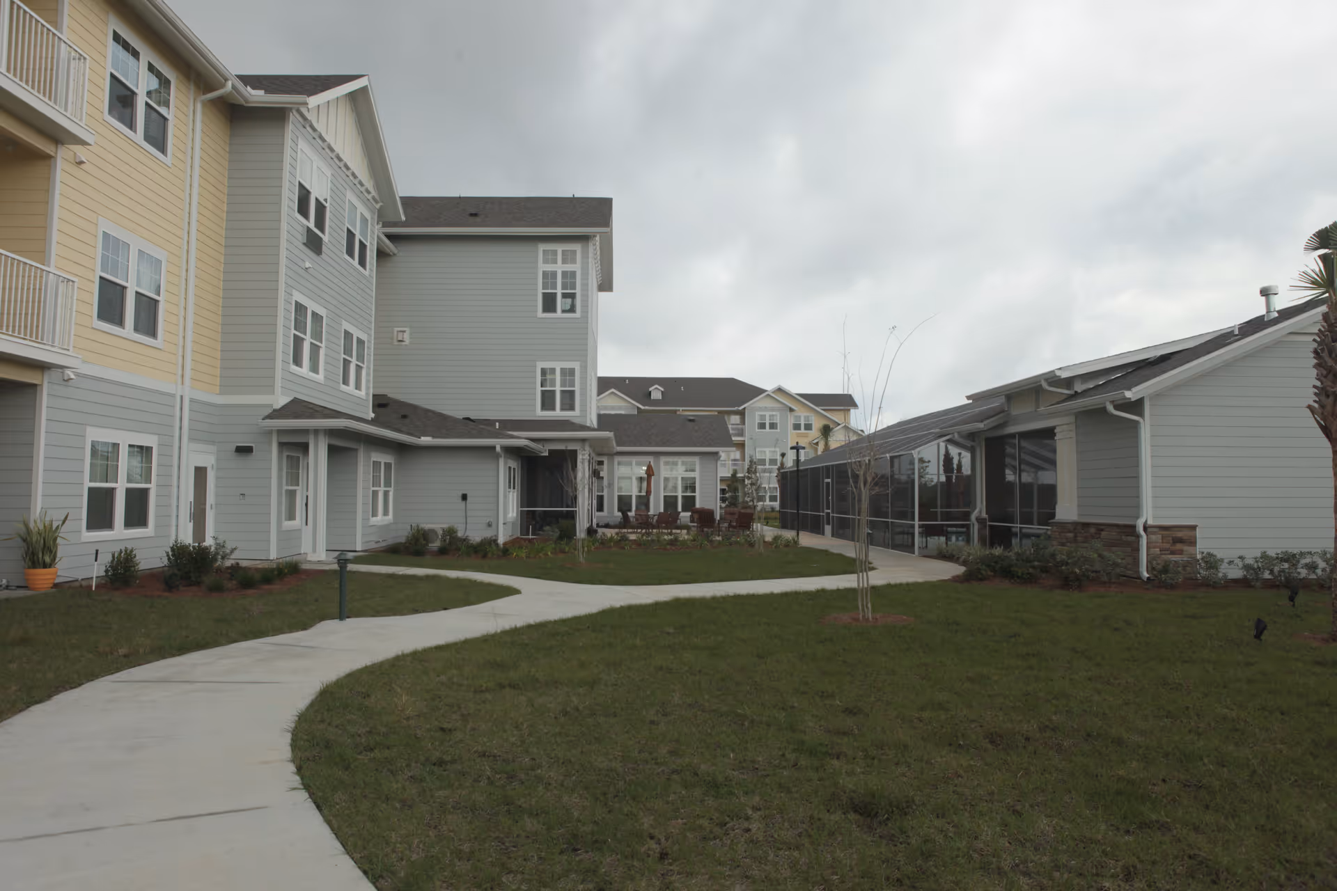 Outdoor view of a senior living facility with a curved concrete walkway, green lawn, and multi-story buildings painted in light yellow and gray. The sky is cloudy and there are some small trees and shrubs planted around the buildings.