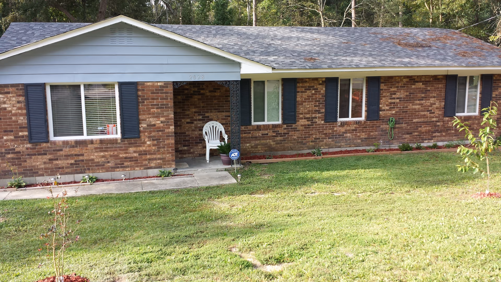 Exterior view of a single-story brick house with a gray shingled roof, several windows with dark shutters, a small covered porch with a white plastic chair and a potted plant, and a well-maintained grassy front yard with small plants and a young tree.