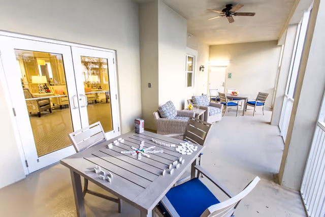 Covered outdoor patio area with a table and chairs, a game of dominoes set up on the table, and additional seating with cushioned armchairs along the wall. The patio is adjacent to a building with glass double doors leading inside.