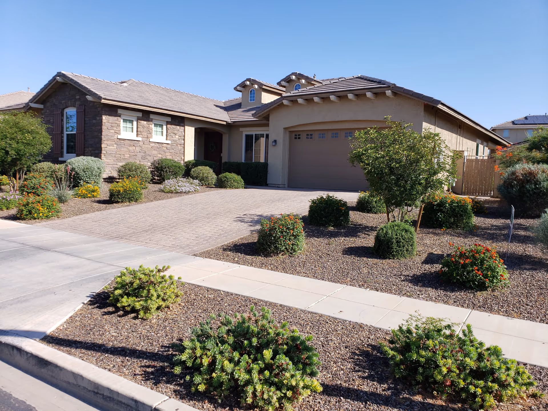 Exterior view of a single-story residential building with a stone and stucco facade, a tiled roof, and a two-car garage. The front yard features desert landscaping with various shrubs, small bushes, and gravel. A concrete sidewalk runs along the front of the property under a clear blue sky.